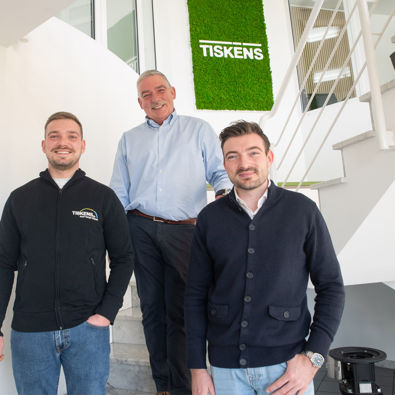 Drei Männer des Tiskens-Teams stehen lächelnd vor weißer Treppe mit grünem Mooswand-Logo im modernen Bürogebäude