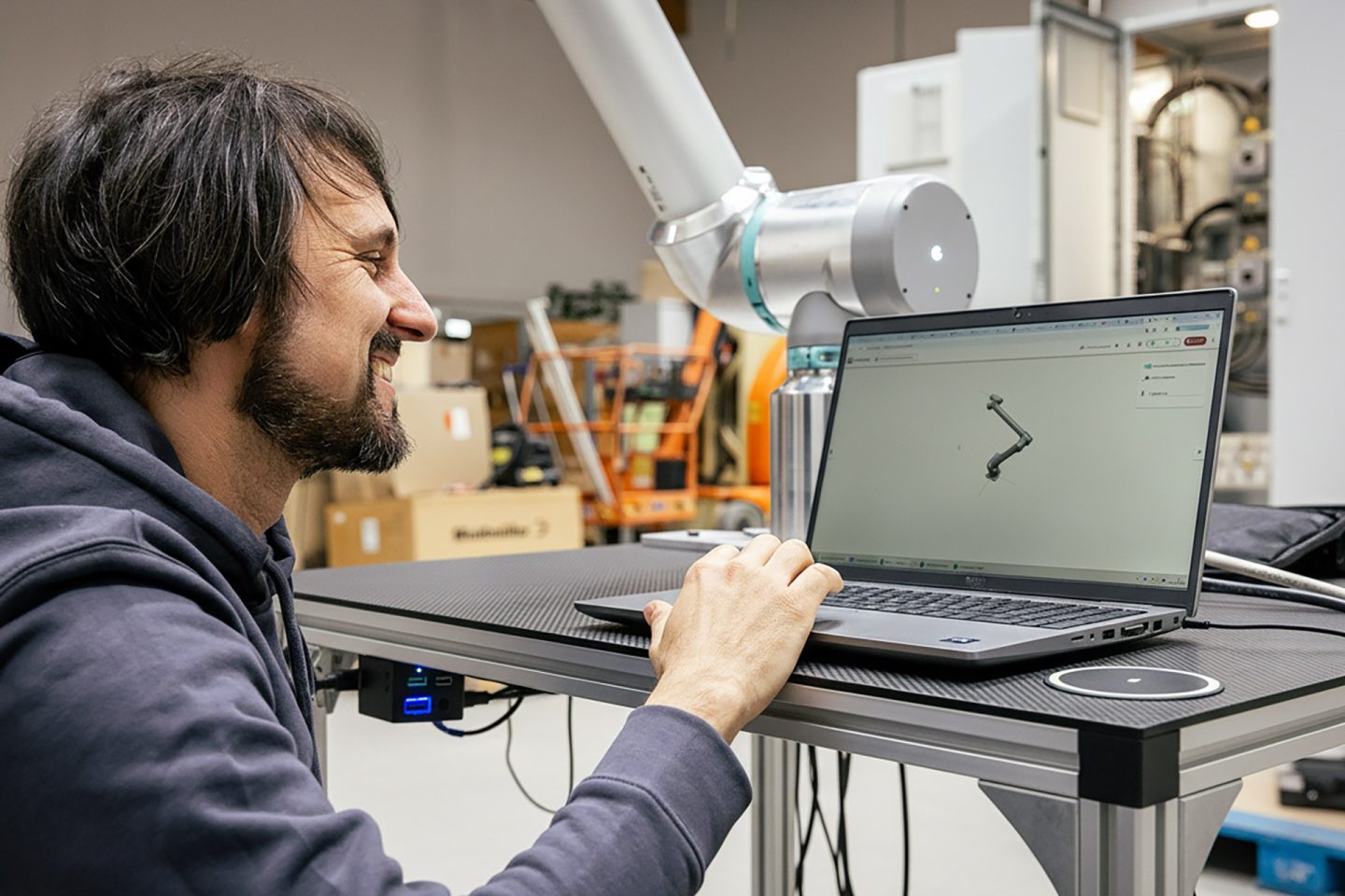 Mann in blauem Hoodie arbeitet am Laptop mit Robotik-Software neben weißem Industrieroboter in moderner Werkstatt