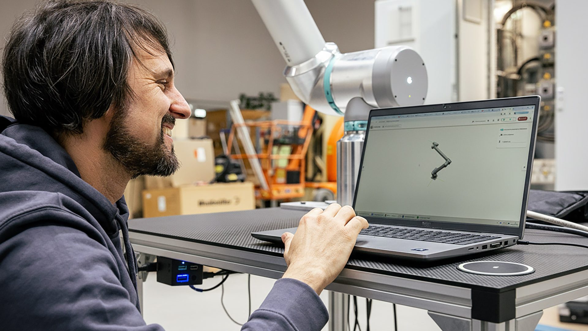 Mann in blauem Hoodie arbeitet am Laptop mit Robotik-Software neben weißem Industrieroboter in moderner Werkstatt