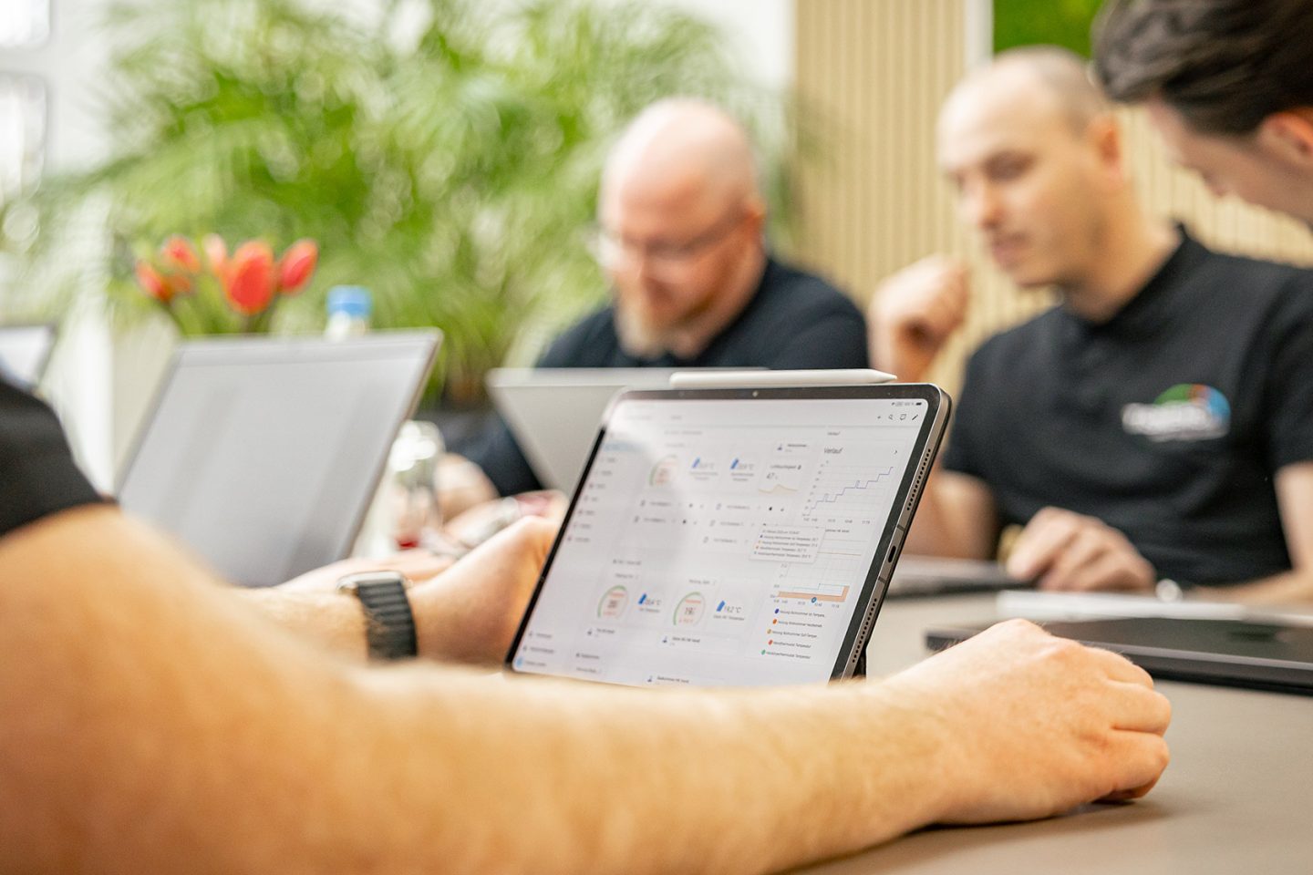 Geschäftsteam arbeitet gemeinsam an Tablets und Laptops in modernem Büro mit Pflanzen im Hintergrund