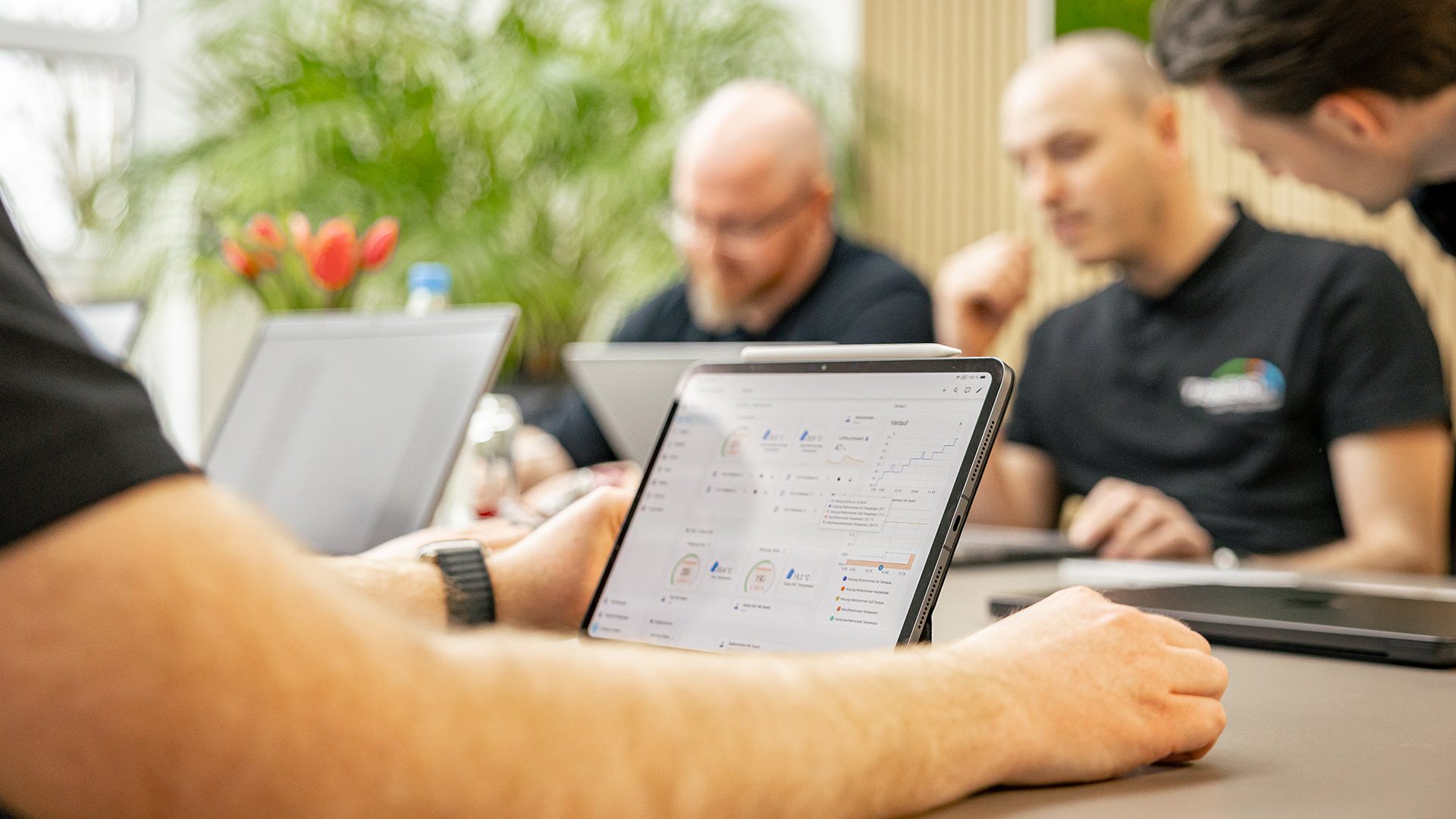 Geschäftsteam arbeitet gemeinsam an Tablets und Laptops in modernem Büro mit Pflanzen im Hintergrund