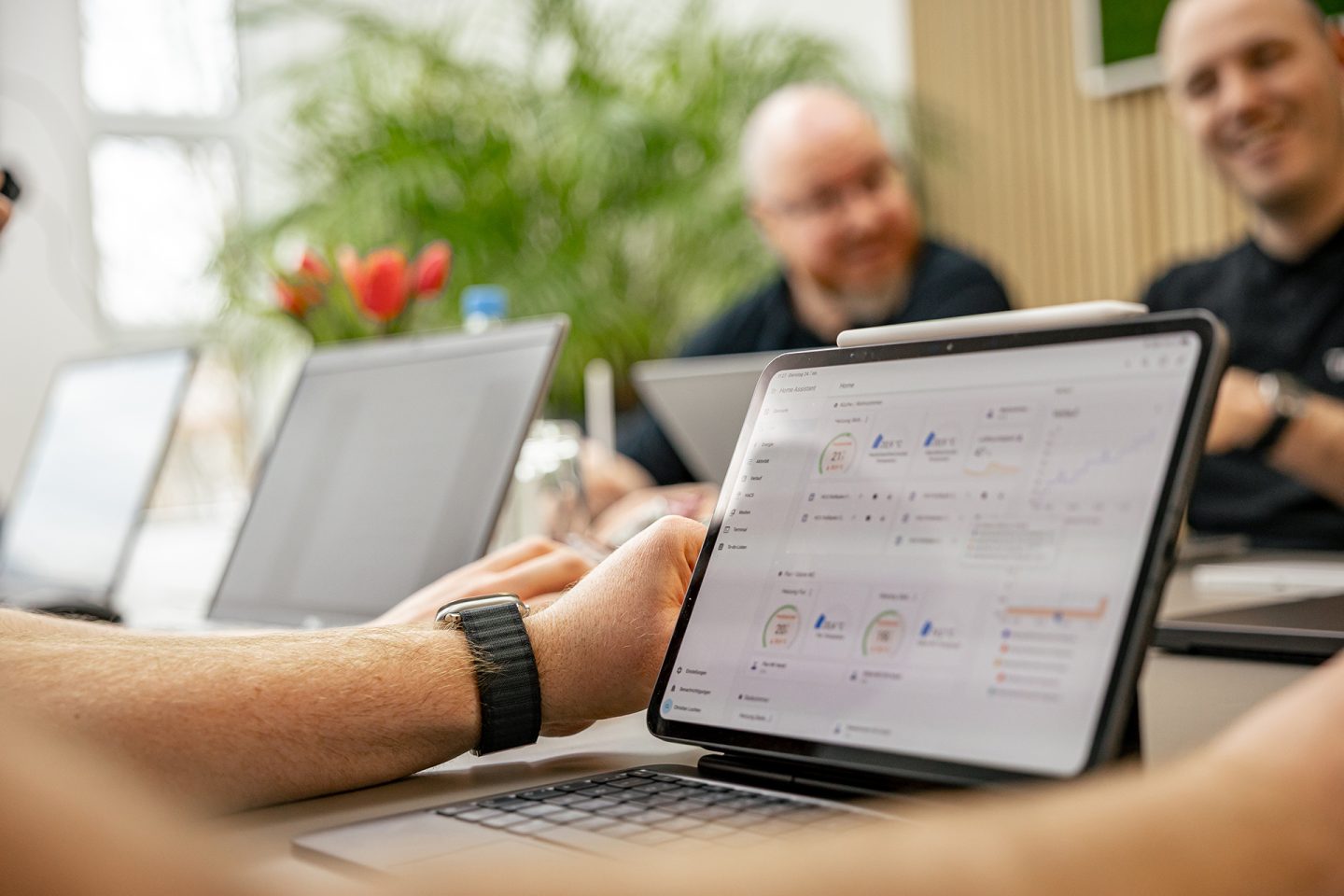 Person mit Smartwatch arbeitet an Laptop mit Dashboard-Ansicht, Kollegen im Hintergrund in hellem Büro