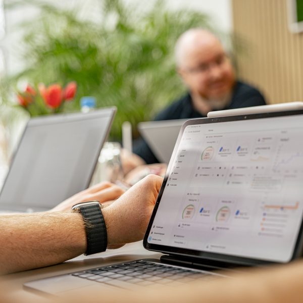 Person mit Smartwatch arbeitet an Laptop mit Dashboard-Ansicht, Kollegen im Hintergrund in hellem Büro