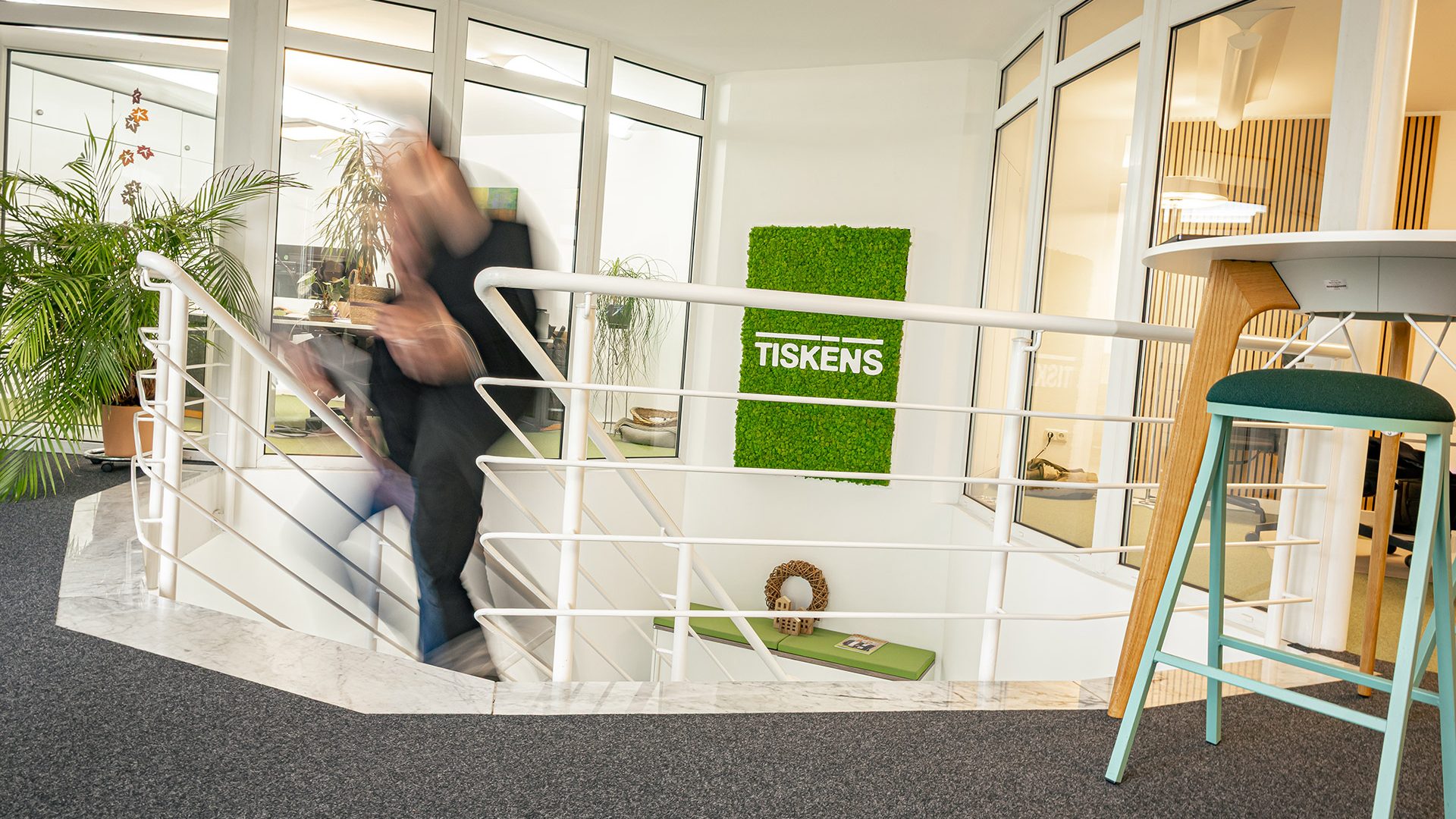 Person läuft Treppe in modernem TISKENS-Büro hinauf, grünes Firmenlogo an Wand, Holzregale und Barhocker sichtbar