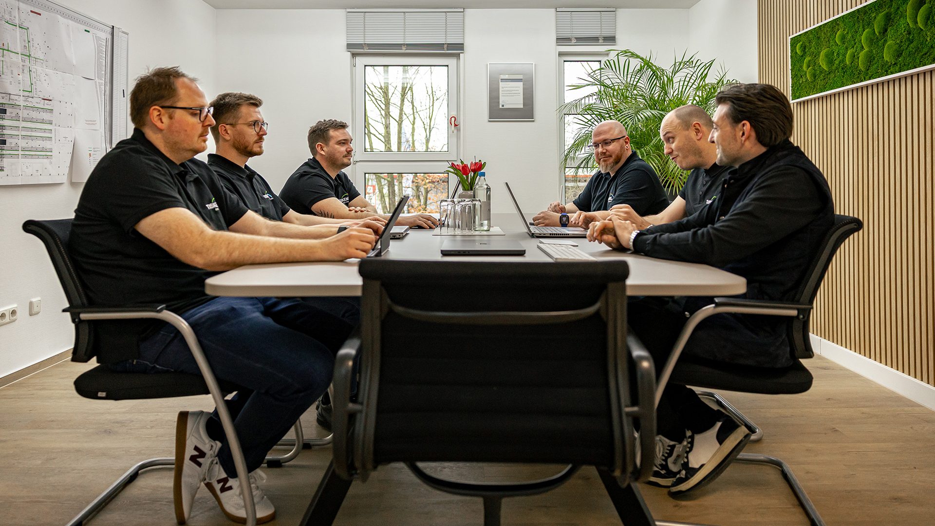 Sechs Männer in schwarzen Poloshirts bei Teambesprechung am modernen Konferenztisch in hellem Büroraum mit Holzwand