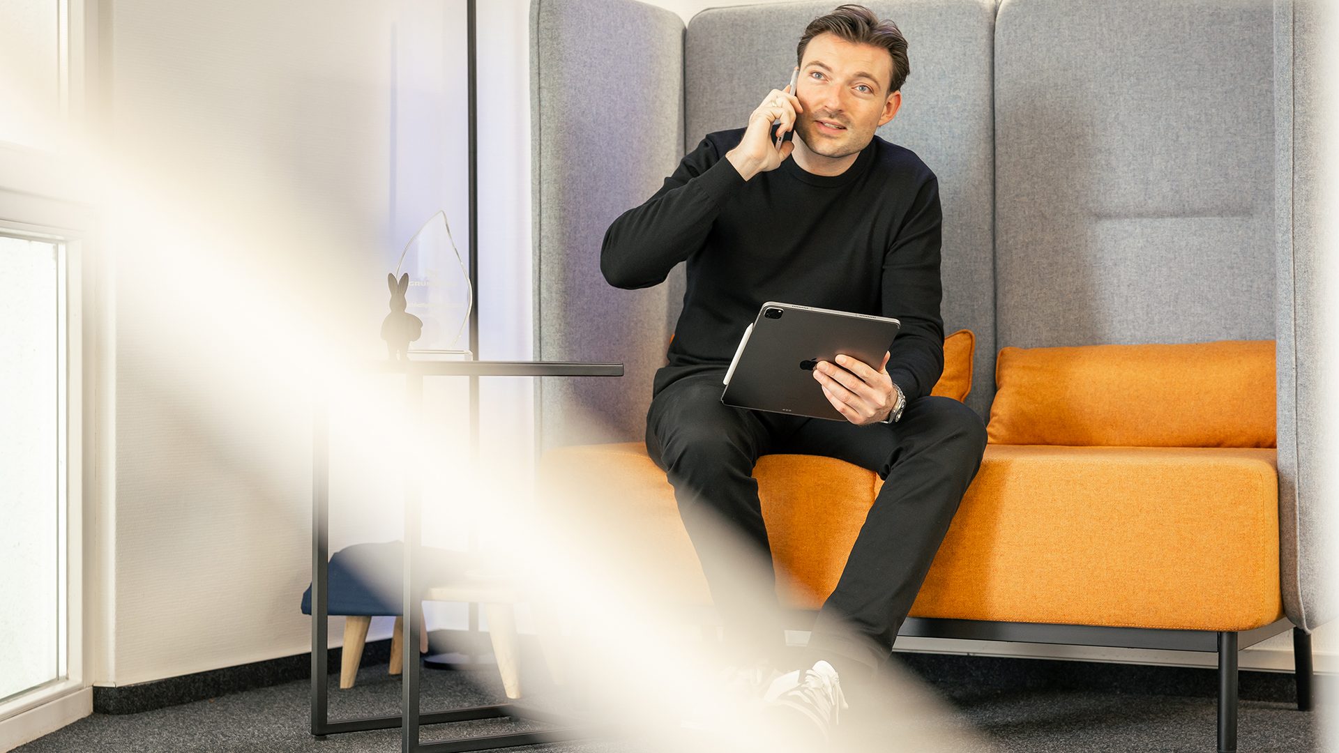 Geschäftsmann telefoniert mit Tablet auf orange-grauem Sessel in modernem Büro