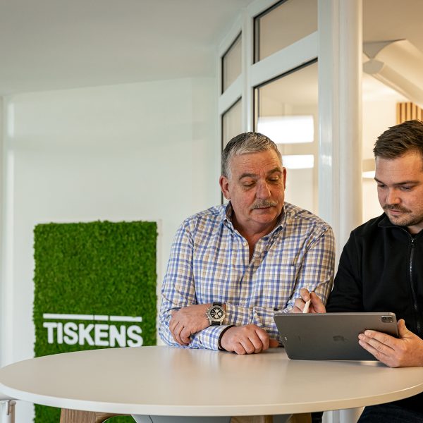 Zwei Männer besprechen Tablet-Inhalte am runden Tisch im modernen Büro mit TISKENS-Logo an grüner Mooswand
