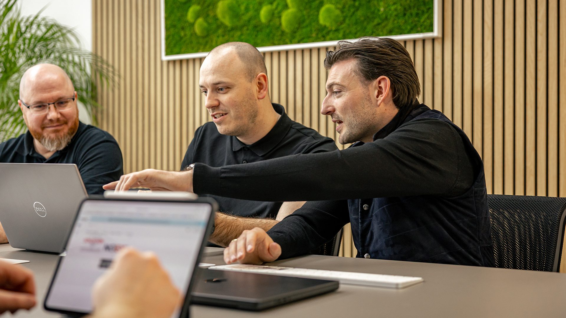 Drei Männer in schwarzer Kleidung arbeiten gemeinsam an Laptops in einem modernen Büro mit Holzwänden