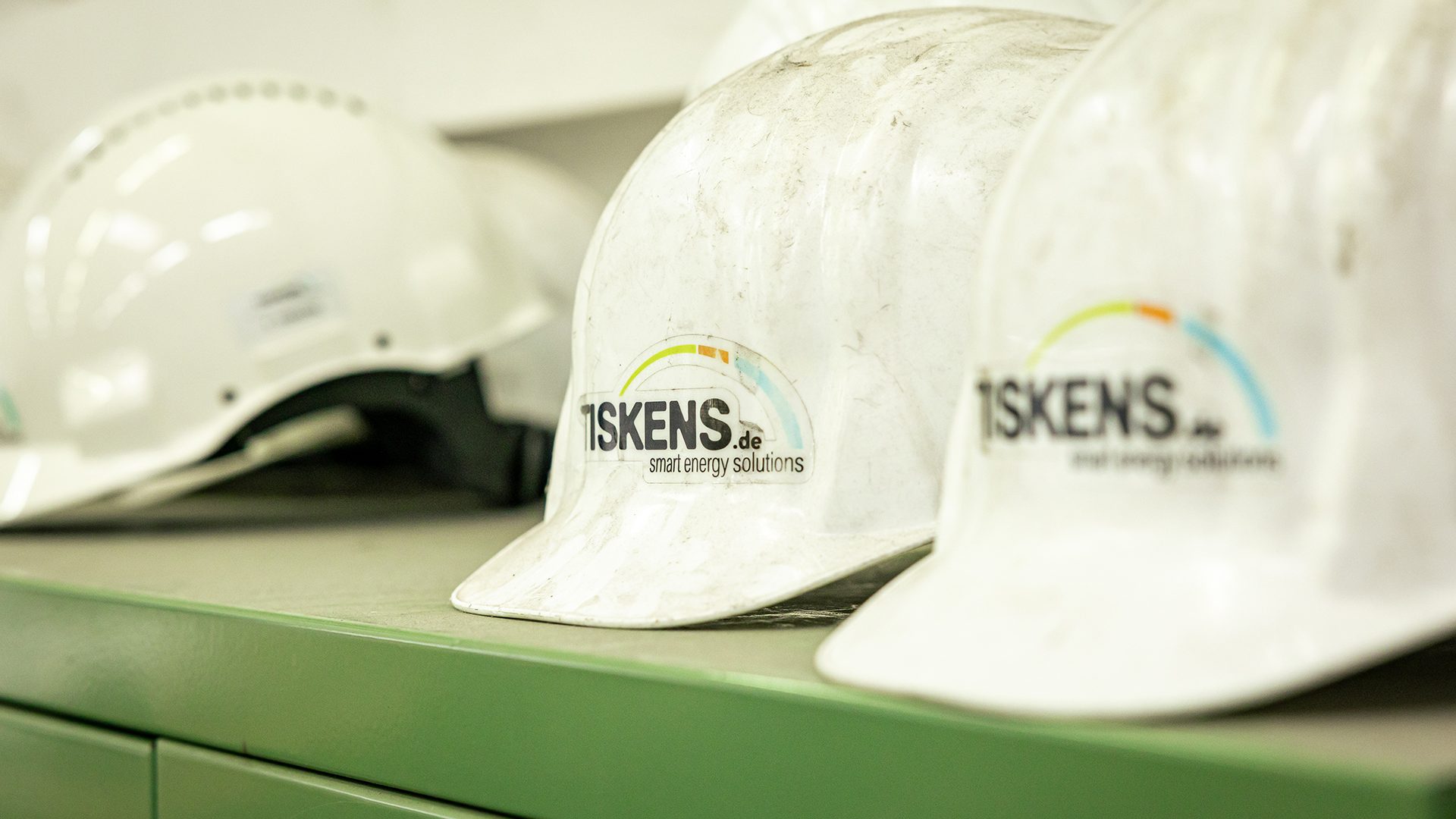 Weiße Bauhelme mit TISKENS Logo und Schriftzug "smart energy solutions" auf grünem Schreibtisch im Büro