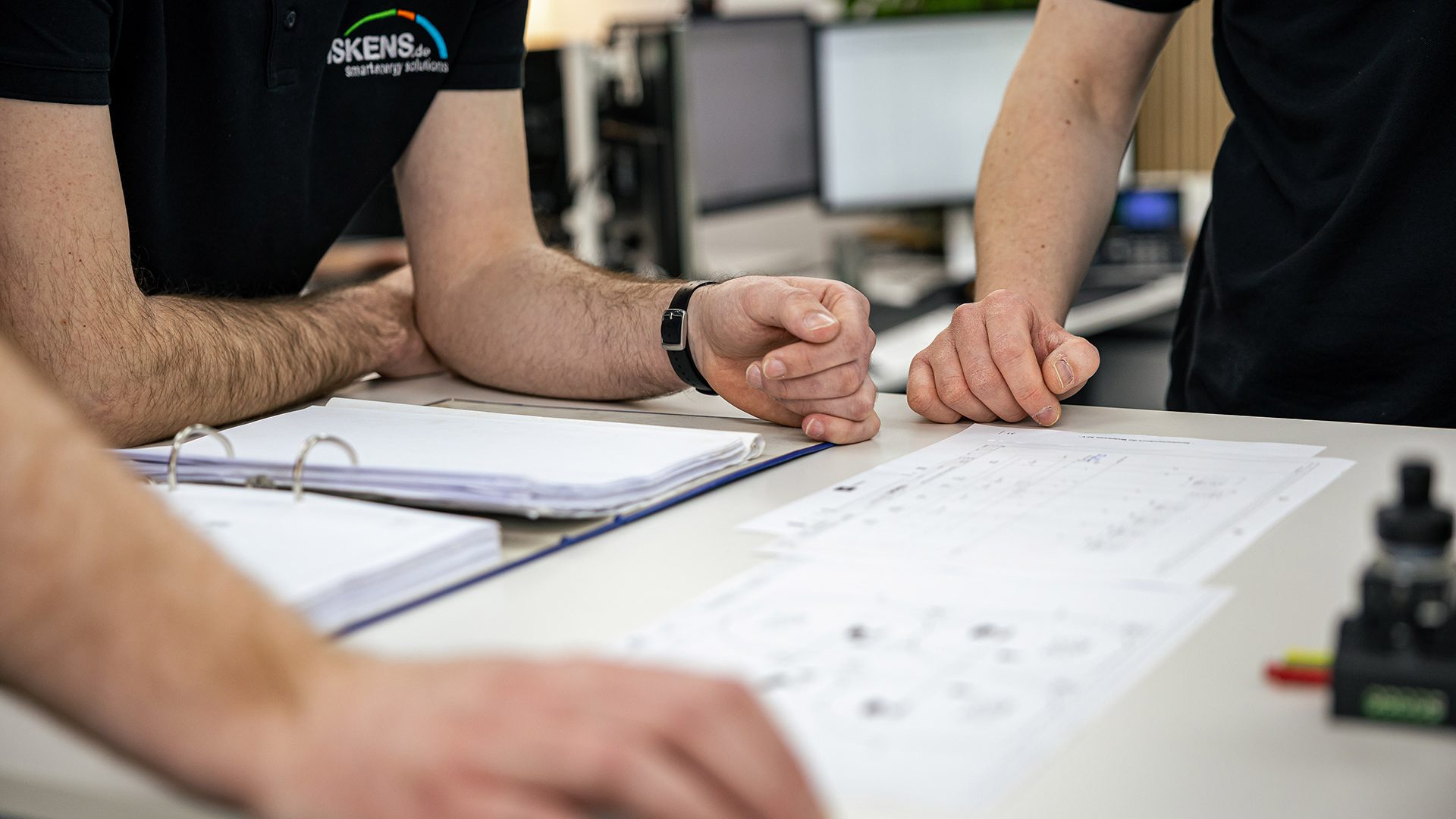 Zwei Personen in schwarzen Shirts betrachten technische Pläne und Dokumente auf einem Schreibtisch im Büro