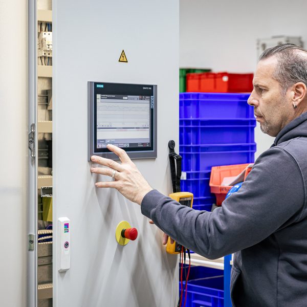 Mann in grauem Kapuzenpullover bedient Touchscreen-Display einer Druckluftanlage in Industriehalle mit bunten Lagerkisten