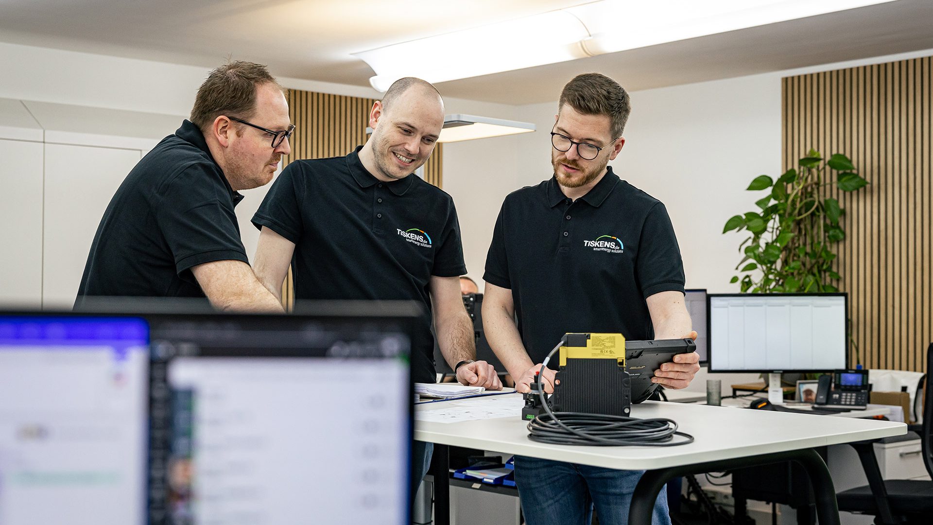 Drei Kollegen in schwarzen Poloshirts analysieren gemeinsam Energiedaten an einem Tablet im modernen Büro