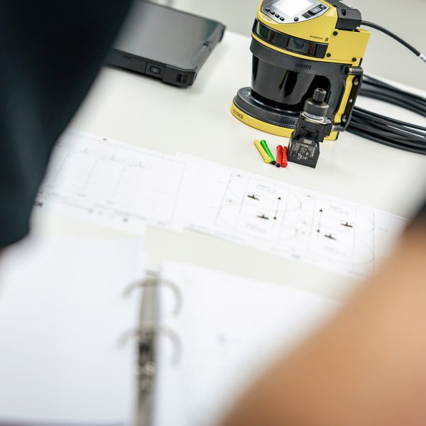 Roboterarm mit gelb-schwarzem Sensor vermisst Bauteile auf weißem Papier neben Laptop und Stift