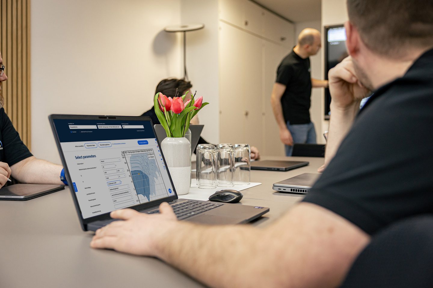 Mann arbeitet am Laptop mit Grundfos-Software zur Pumpenauslegung während Kollegen im Hintergrund diskutieren