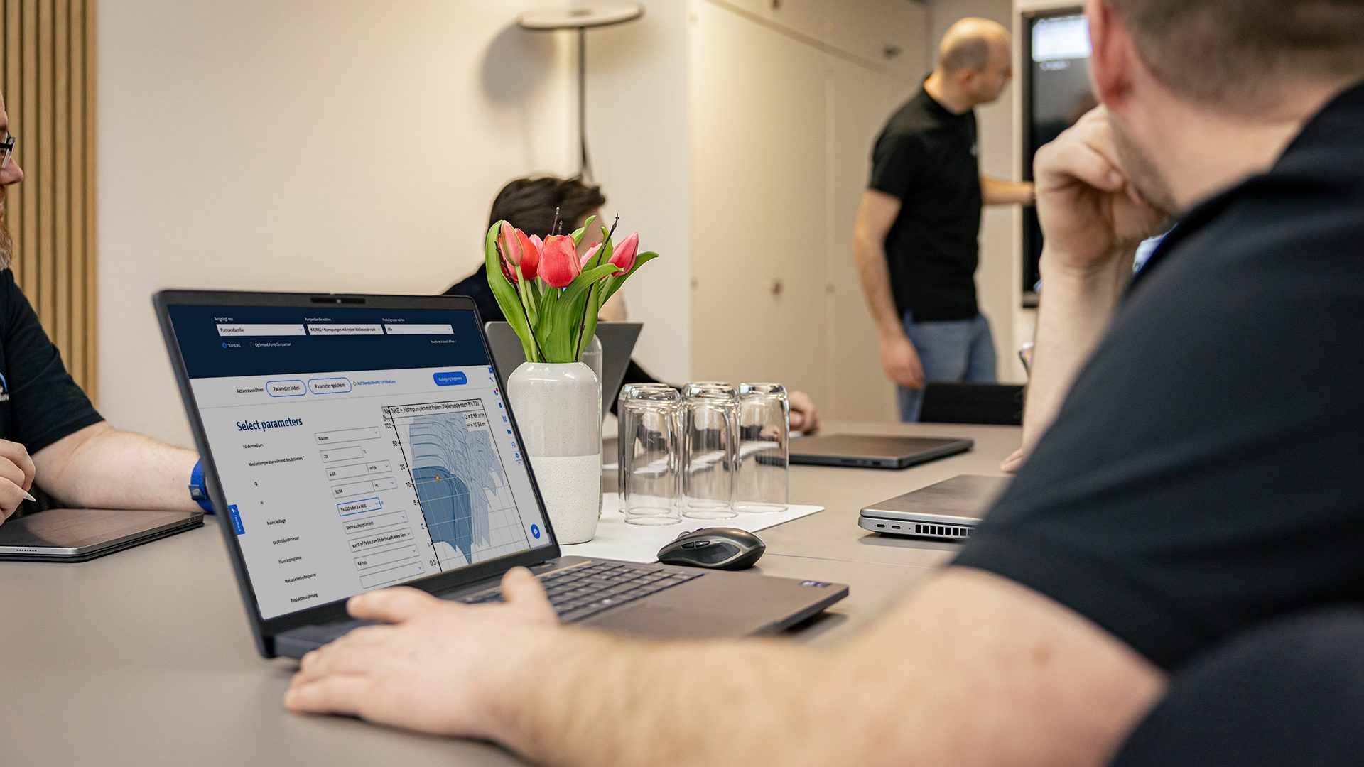 Mann arbeitet am Laptop mit Grundfos-Software zur Pumpenauslegung während Kollegen im Hintergrund diskutieren