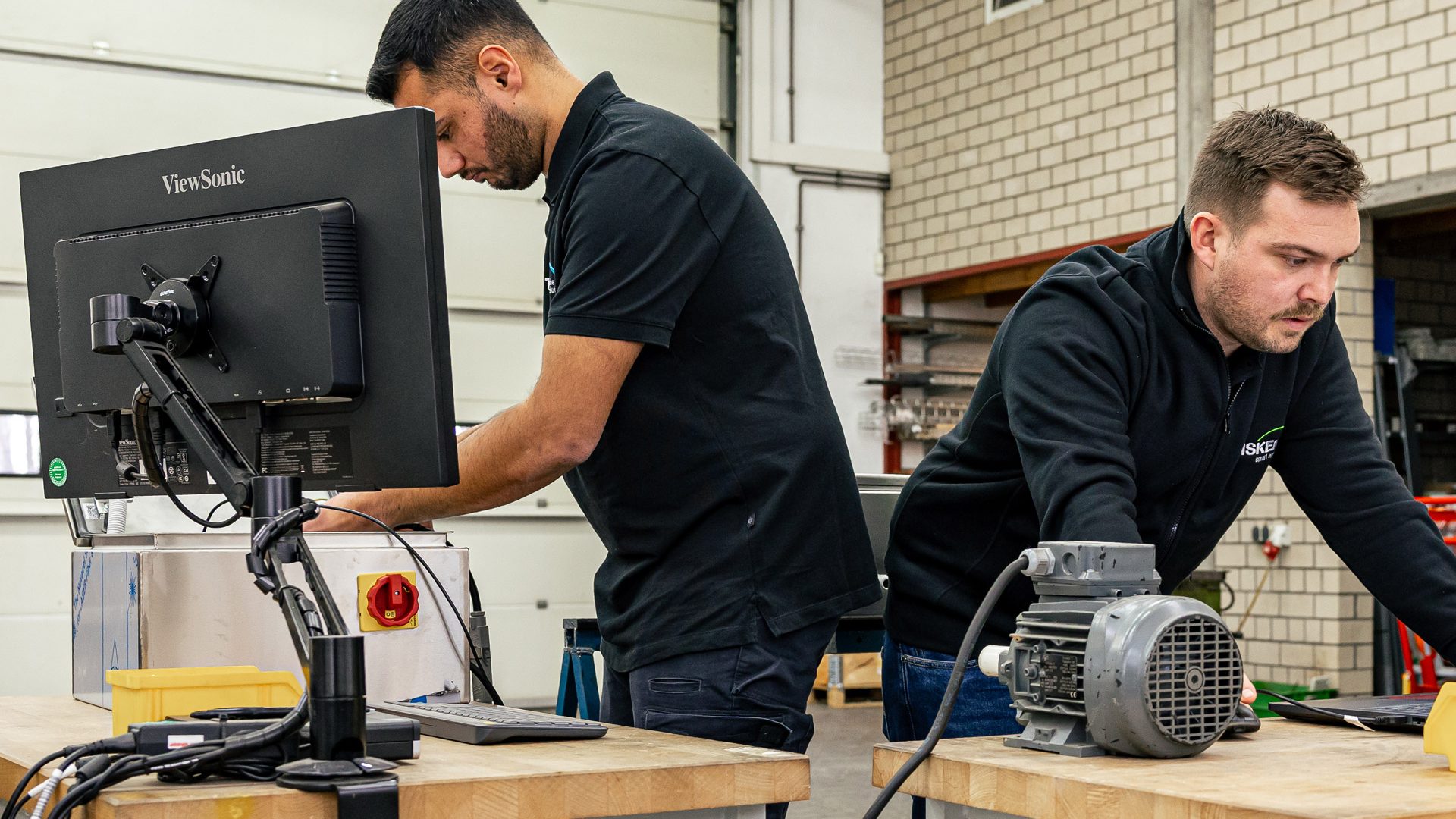 Zwei Techniker arbeiten an Werkbank mit Computer und Elektromotor in industrieller Werkstatt für Retrofit-Modernisierung