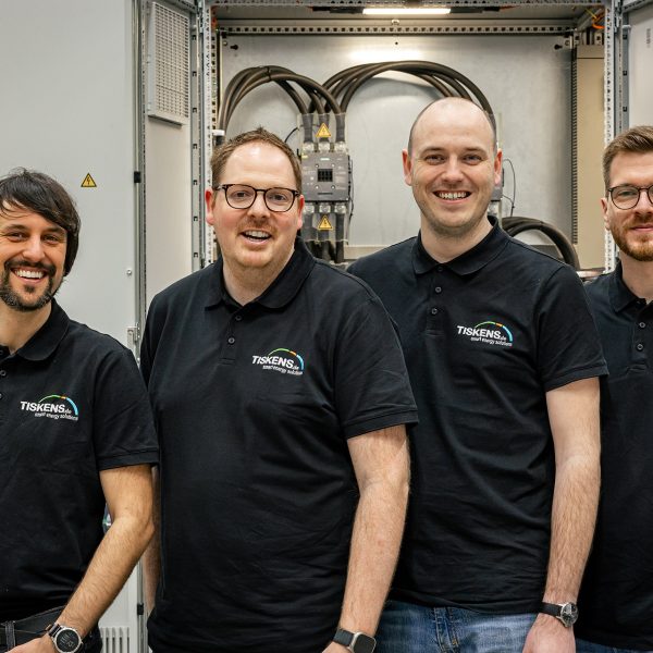 Vier Männer in schwarzen Tiskens-Poloshirts stehen lächelnd vor industrieller Automatisierungsanlage mit Schaltschrank