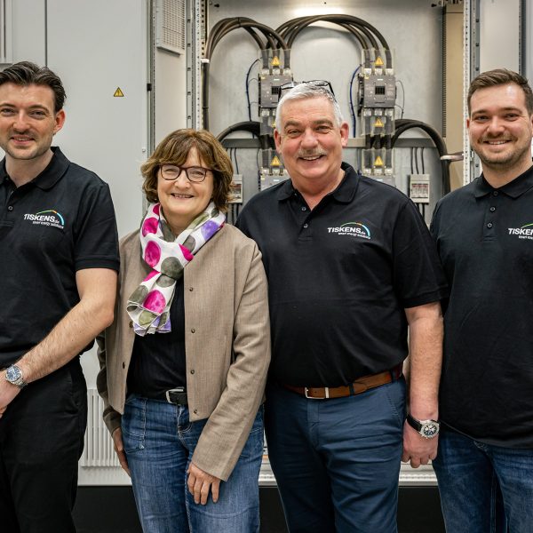 Vier Personen stehen lächelnd vor einer Industriemaschine, drei Männer in schwarzen Tiskens-Poloshirts und eine Frau in