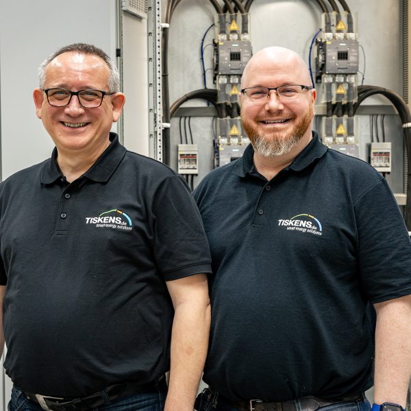 Zwei Techniker in TISKENS-Poloshirts vor elektrischen Schaltanlagen im Servicezentrum